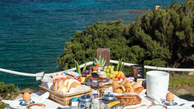 Frühstückstisch auf einer Terrasse mit traumhaftem Blick auf das tiefblaue Meer – serviert werden frische Croissants, Brötchen, Obst, Säfte und Kaffee – ein perfekter Start in den Tag mit mediterranem Panorama.