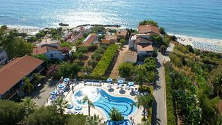 Luftaufnahme eines Hotels an der Küste mit geschwungenem Außenpool, umgeben von Palmen und Sonnenschirmen; im Hintergrund das tiefblaue Meer und ein Sandstrand mit Liegen.