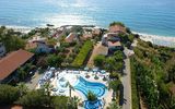 Luftaufnahme eines Hotels an der Küste mit geschwungenem Außenpool, umgeben von Palmen und Sonnenschirmen; im Hintergrund das tiefblaue Meer und ein Sandstrand mit Liegen.