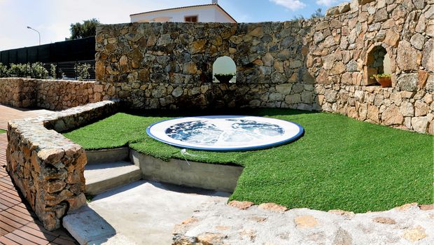 Kleiner Whirlpool im Freien, eingebettet in eine charmante Steinmauer mit rustikalem Flair – umgeben von Kunstrasen und mediterranem Ambiente für entspannte Momente unter freiem Himmel.