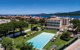 Luftaufnahme eines stilvollen Hotels mit großem Außenpool inmitten gepflegter Gartenanlagen – mit Blick auf die umliegenden Häuser, das tiefblaue Meer und die grünen Hügel Sardiniens.