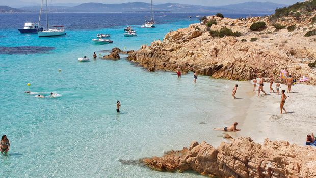 Traumhafter Strand mit türkisfarbenem Wasser, weißen Sandbuchten und badenden Urlaubern – umgeben von Felsen und Booten vor malerischer Küstenkulisse.