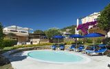 Kleiner Rundpool mit blauen Sonnenschirmen und Liegestühlen, umgeben von gepflegten Gärten und einer mediterranen Hotelanlage mit blühender Bougainvillea unter strahlend blauem Himmel.