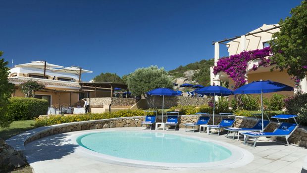 Kleiner Rundpool mit blauen Sonnenschirmen und Liegestühlen, umgeben von gepflegten Gärten und einer mediterranen Hotelanlage mit blühender Bougainvillea unter strahlend blauem Himmel.
