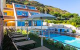 Moderne Hotelanlage mit bunten Fassaden, sonniger Liegewiese und großzügigem Poolbereich inmitten grüner Hügellandschaft.