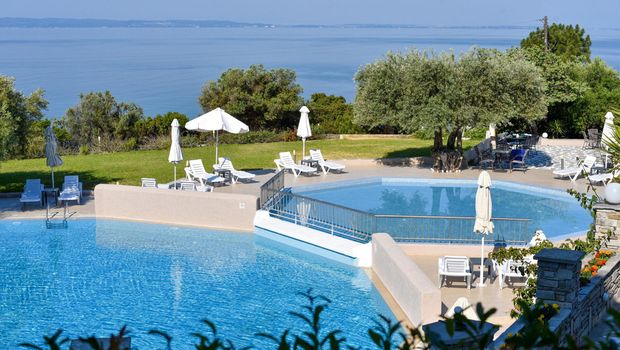 Zwei miteinander verbundene Außenpools mit Panorama-Meerblick, umgeben von Sonnenliegen, Olivenbäumen und mediterranem Garten – ein Ort zum Entspannen und Genießen.