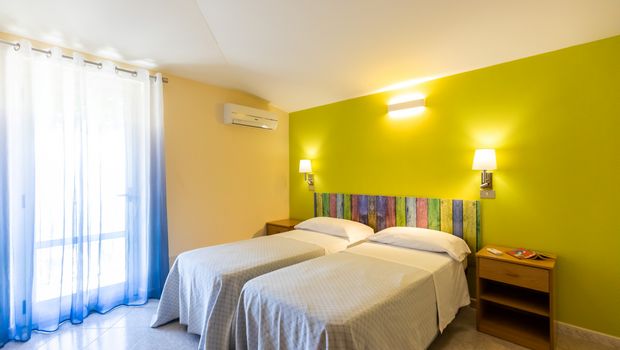 Helles Hotelzimmer mit zwei getrennten Einzelbetten, farbigem Holz-Kopfteil und gelbgrüner Wand; links ein Fenster mit halbtransparentem Vorhang in Blau-Weiß-Verlauf.