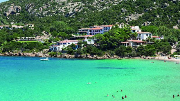 Idyllische Bucht mit kristallklarem, türkisblauem Wasser – im Hintergrund eingebettet in grüne Hügel liegen charmante weiße Hotelgebäude mit roten Dächern, während Badegäste das Meer genießen.